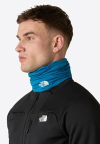 Turkos halsduk av elastiskt stoff, med en vit logotyp. Bärs över en svart zip-up jacka. Slät textur, modern design.