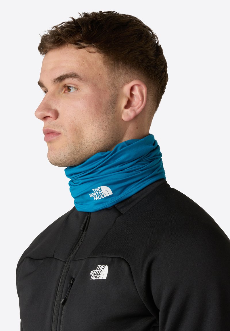 Turkos halsduk av elastiskt stoff, med en vit logotyp. Bärs över en svart zip-up jacka. Slät textur, modern design.