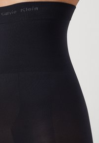 Black sheer tights with a high-waisted, ribbed waistband featuring subtle branding. Smooth texture with a regular fit.