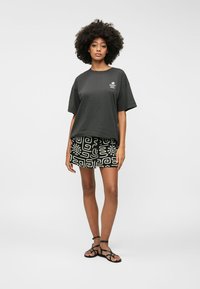 Camiseta de algodón gris con corte holgado y logo en el pecho, combinada con pantalones cortos negros con estampado que presenta diseños geométricos blancos y sandalias planas.