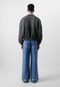 Sweatshirt oversized gris foncé avec épaules tombantes, associé à un jean large bleu avec des accents déchirés et des éclaboussures de peinture blanche.