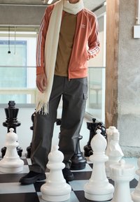 Orange atletisk jacka med vita ränder, brun skjorta, grå cargobyxor och en krämfärgad scarf. Står bland stora schackpjäser i svart och vitt.