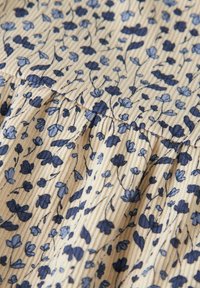 Lichtbeige stof met verticale ribbeling heeft een blauw bloemmotief, inclusief kleine bloesems en bladeren, wat een textuur aan het geheel geeft.