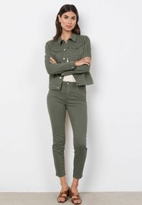 Olijfgroene denimjacket met knoopsluitingen en een gestructureerde kraag, gecombineerd met bijpassende strakke olijfgroene broek en bruine sleehakken.