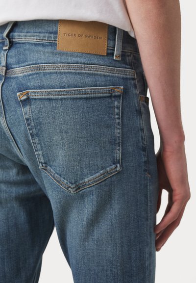 Jean en denim bleu avec une étiquette en cuir beige portant la mention "TIGER OF SWEDEN", quatre poches et des surpiqûres orange au dos.