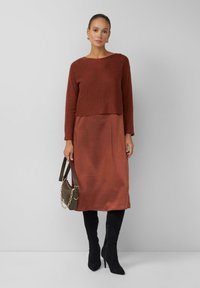 Frau in einem rostfarbenen Strickpullover und passendem Midikleid, die eine braune Handtasche mit goldener Kette und schwarze kniehohe Absatzstiefel trägt.