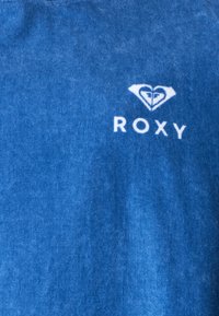 Μπλε ύφασμα με λευκό λογότυπο της μάρκας Roxy που απεικονίζει στυλιζαρισμένη καρδιά και κύμα πάνω από το κείμενο "ROXY" σε υφασμάτινη επιφάνεια με υφή.