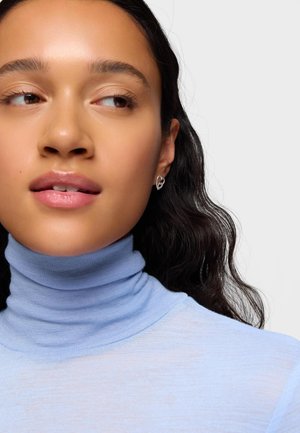 Joven con cabello largo y oscuro, lleva un suéter de cuello alto azul claro y pendientes plateados en forma de corazón, mirando ligeramente hacia un lado.