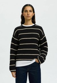 Pullover nero a maglia con strisce orizzontali bianche, dotato di un collo rotondo e maniche lunghe. Indossato sopra una maglietta bianca a strati, abbinato a jeans blu.
