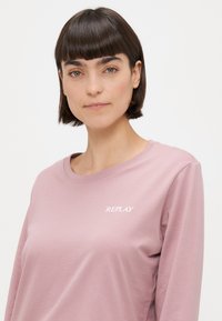 Koszula z długim rękawem w jasnoróżowym kolorze, wykonana z miękkiego materiału. Posiada okrągły dekolt oraz białe logo "REPLAY" na piersi. Prosty design.