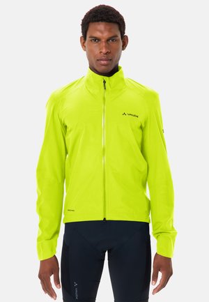 Man wearing bright yellow zip-up cycling jacket and black cycling shorts, standing against plain white background.