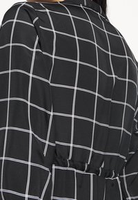 Černý oděv s bílým rastrovým vzorem. Texturovaná tkanina s elastickým pásem a přizpůsobeným střihem na zádech.