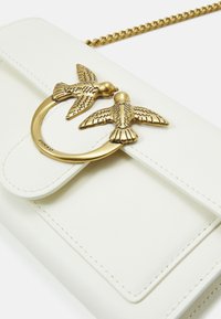 Bijela torba od umjetne kože za crossbody nošenje s zlatnim dvostrukim kopčama u obliku ptica, okruglim hardverom i ušivenim detaljima. Ima lančanu naramenicu.