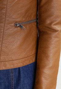 Détail de la manche de veste en cuir marron et de la poche zippée sur un jean en denim bleu, mettant en évidence la texture et la couture.