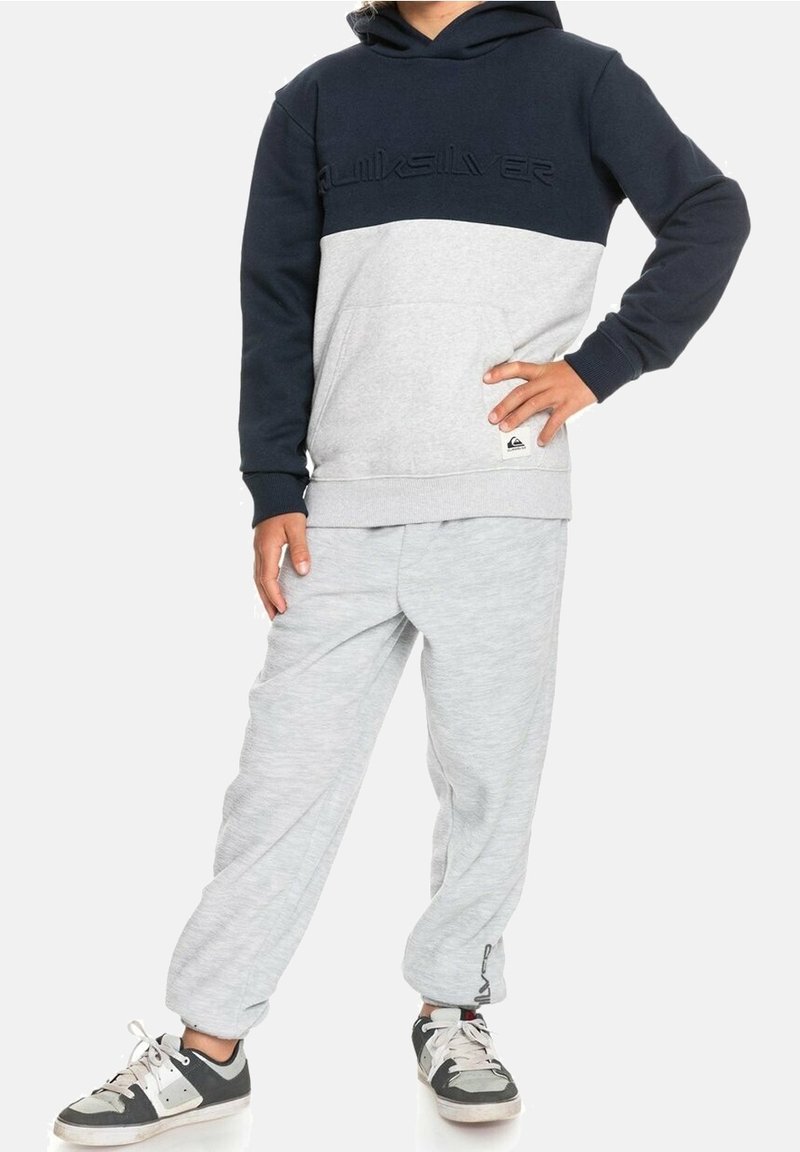 Enfant portant un sweat à capuche Quiksilver bleu marine et gris, un pantalon de jogging gris clair, et des baskets noires et blanches, debout avec une main sur la hanche.