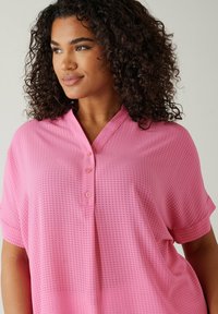 Blusa rosa a maniche corte con finitura texture, dotata di una patta con bottoni e una vestibilità rilassata. Tessuto leggero con un sottile motivo a quadri.