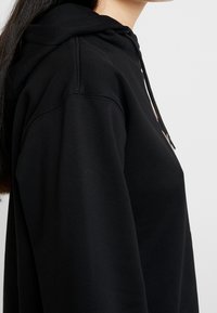 Vista lateral de una persona con una sudadera negra y cabello largo y oscuro contra un fondo claro y liso.