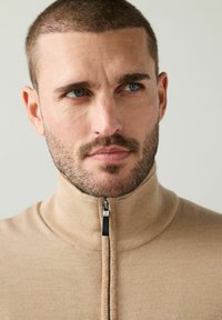 Šviesiai rudas zip-up švarkelis su aukštu apykakle, tekstūruota medžiaga ir juoda užtrauktuko detalė. Artimas kadras, kuriame matoma apykaklės zona ir veido bruožai.