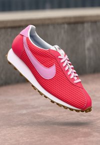 Sapatilha de corrida Nike em vermelho e rosa com atacadores brancos e sola de borracha, mostrada em ângulo acima de uma superfície de pedra e com fundo desfocado.
