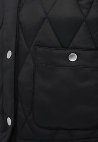 Μαύρο γιλέκο με καπιτονέ σχέδιο διαγώνιας ραφής, με εμπρόσθια τσέπη που κλείνει με ασημί κουμπί και λαμπερές μεταλλικές λεπτομέρειες.