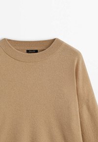 Beige stickad tröja med ribbad rund halsringning, sänkt axelsöm och mjuk textur. Etikett synlig vid halsområdet.