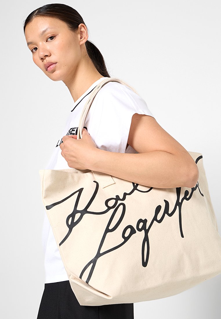 Jeune femme aux cheveux noirs attachés qui porte un grand sac en toile beige avec un texte en noir audacieux sur l'épaule, portant un haut sans manches blanc.