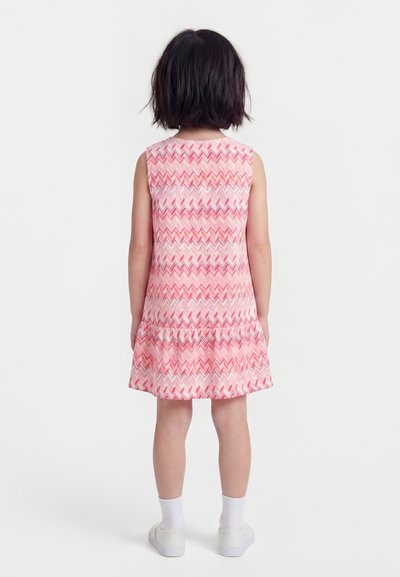 Παιδί με κοντά σκούρα μαλλιά στέκεται γυρίζοντας προς τα πίσω, φοράει αμάνικο ροζ φόρεμα με σχέδιο ζιγκ-ζαγκ, λευκές κάλτσες και λευκά αθλητικά παπούτσια.