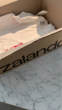 Zalando kartonska škatla z debelimi črnimi črkami, ki vsebuje zmečkani bel srajco z rdečo etiketo. Površina je z vzorcem marmorja.