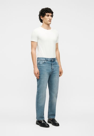 Άνδρας με σγουρά σκούρα μαλλιά που φοράει απλό λευκό μπλουζάκι, ανοιχτό μπλε τζιν και μαύρα δερμάτινα παπούτσια, στέκεται μπροστά σε λευκό φόντο.