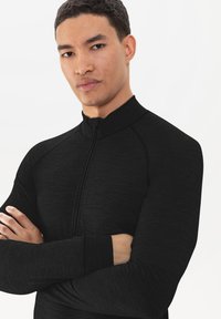 Zwart sportief zip-up top, textielstof, hoge kraag, lange mouwen met boorden. Model heeft de armen gekruist, wat de pasvorm van het kledingstuk toont.