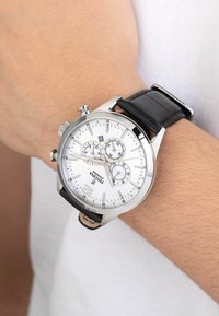 Sidabrinis chronograph laikrodis su baltu ciferblatu, juodu odiniu dirželiu, trimis subdiais, datos rodikliu ir poliruotu metaliniu korpusu, uždėtas ant riešo.
