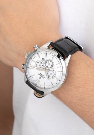 Montre chronographe argentée avec un cadran blanc, un bracelet en cuir noir, trois sous-cadrans, un affichage de la date et un boîtier en métal poli, portée au poignet.