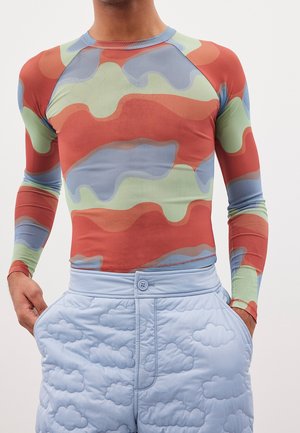 Man wearing fitted long-sleeve top with multicolor abstract pattern and light blue quilted pants with stitched cloud designs, hands in pockets.
