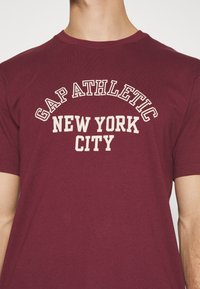 Camiseta marón de algodón, con "GAP ATHLETIC NEW YORK CITY" impreso en blanco en un diseño arqueado en el pecho. Mangas cortas.
