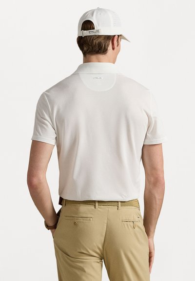 Polo em branco com mangas curtas, apresentando um colarinho e um logotipo nas costas. Combinado com calças bege e um boné correspondente.