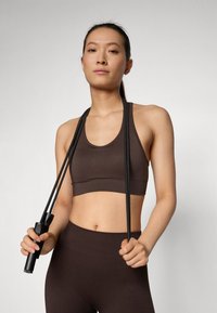 Soutien-gorge de sport marron avec une texture côtelée, associé à un legging taille haute assorti, tenant une corde à sauter noire avec des poignées texturées.