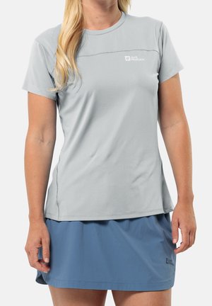 Kobieta w jasnoszarej koszulce z krótkim rękawem i niebieskiej spódnicy, stojąca, z blond włosami opadającymi na jedno ramię.