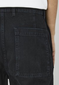 Pantalons en denim noir avec une poche arrière et des coutures visibles. Comprennent une taille standard avec passants de ceinture, affichant une texture robuste.