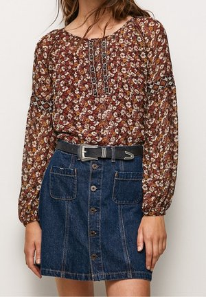 Kobieta nosząca kwiecistą, długorękawową bluzkę wpuszczoną w spódnicę jeansową z wysokim stanem, zapinaną na guziki z naszywanymi kieszeniami oraz czarny pasek z srebrną klamrą.