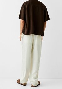 Osoba stojící zády k bílé zdi, oblečená v bílých volných kalhotách, hnědém krátkém triku a hnědých semišových nazouvacích botách.