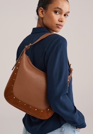 Sac à bandoulière en cuir marron avec accents en daim et détails métalliques cloutés. La forme incurvée comprend une bandoulière réglable unique pour un port facile.