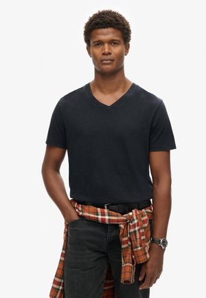 Camiseta negra de cuello en V, de mangas cortas. Jeans de mezclilla oscuros con bolsillos. Camisa de cuadro naranja atada a la cintura. Reloj plateado en la muñeca.