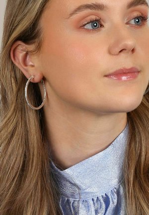 Silverfärgade ringörhängen med en texturerad design, som bärs av en modell och kompletterar en ljusblå, glänsande, kragad topp.