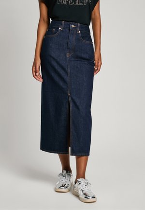 Femme portant une jupe midi en jean bleu foncé avec une fente devant et des baskets blanches, debout devant un fond clair uni.