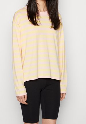 Frau trägt ein langärmeliges Hemd mit pastellgelben und rosa Streifen und schwarze Radhosen vor einem schlichten Hintergrund.