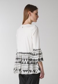 Vit l�ångärmad topp med en svart mönstrad peplumfåll och trumpetärmar. Har en rundad halsringning och en liten öppning bak.