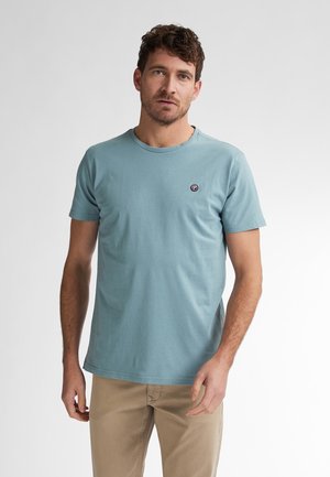 Camiseta de algodón azul claro con mangas cortas, cuello redondo y pequeño logo circular en el lado izquierdo del pecho. Textura suave, corte casual.