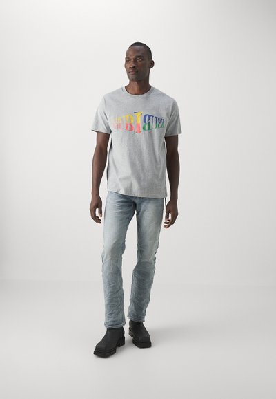 Grå bomulls-T-shirt med färggrafik, tillsammans med ljusblå jeans och svarta ankelstövlar. Enkel rund halsringning och korta ärmar.