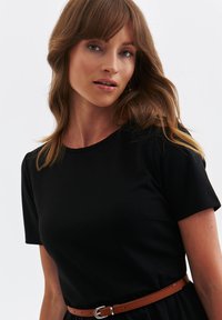 Kobieta z jasnobrązowymi falującymi włosami, ubrana w czarny t-shirt z krótkim rękawem oraz cienki brązowy pasek, na jednolitym jasnym tle.