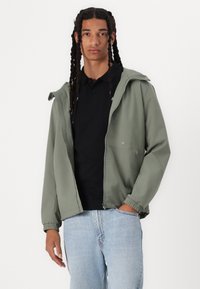 Jachetă verde deschis cu glugă, prevăzută cu închidere cu fermoar și manșete elastice. Purtată peste un tricou polo negru, stilizată cu blugi albaștri.